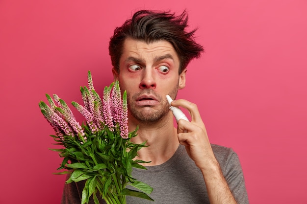 Shocked sick man holds nasal drops, has watery red eyes, suffers from allergy on pollen, has inflammation of nose, reacts on environmental allergen, has very sensitive immune system. Hay fever