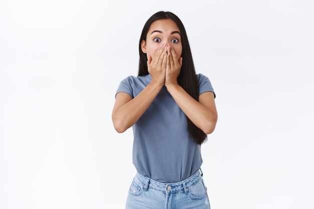 Shocked and speechless emotional asian woman witness something terrible, popping eyes, gasping, hold hands on mouth not scream in panic, standing white wall astonished or terrified