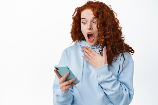 Shocked and startled redhead girl scream while reading message on smartphone, staring at mobile screen with surprised face, standing  on white