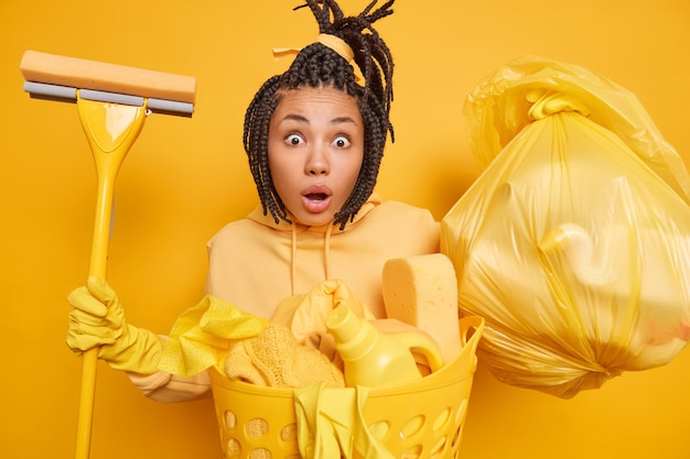 Free photo shocked surprised ethnic housemaid wears rubber gloves for hand protection holds mop trash bag