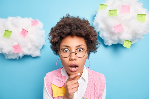 Free photo shocked terrified female student uses important note uses paper stickers post keeps hand on chin being deeply surprised wears round spectacles formal clothes isolated over blue wall