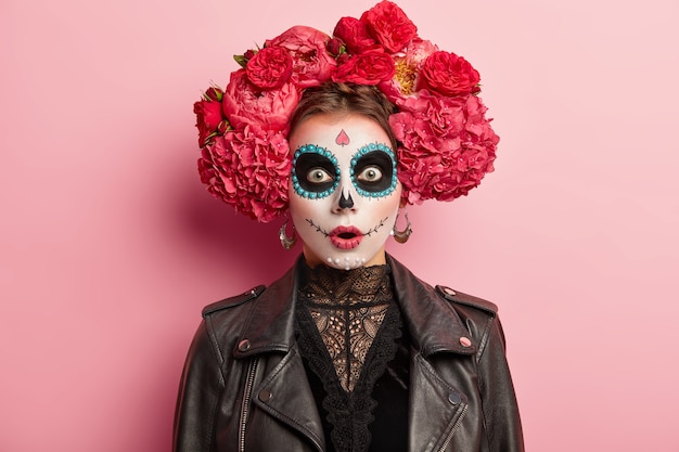 Shocked terrified young woman has scary ghost face, wears artistic makeup for Day of Dead holiday, wears black leather jacket, models over rosy studio background. Skull female symbolizing death