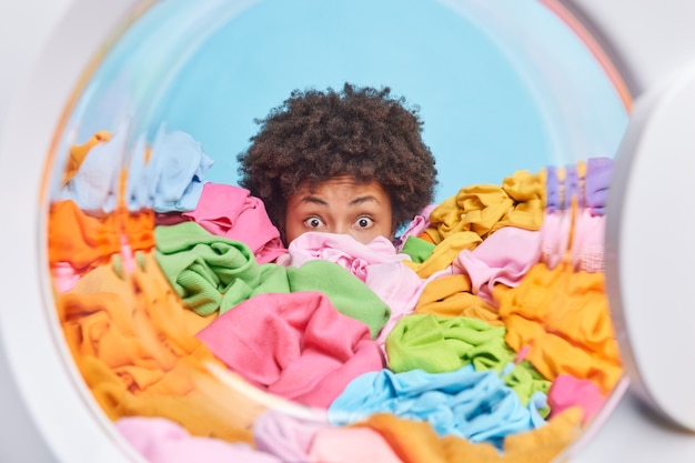 Free photo shocked troubled woman hides behind big pile of laundry oveloaded with housework and domestic responsibilities stares bugged eyes poses from washing machine drum