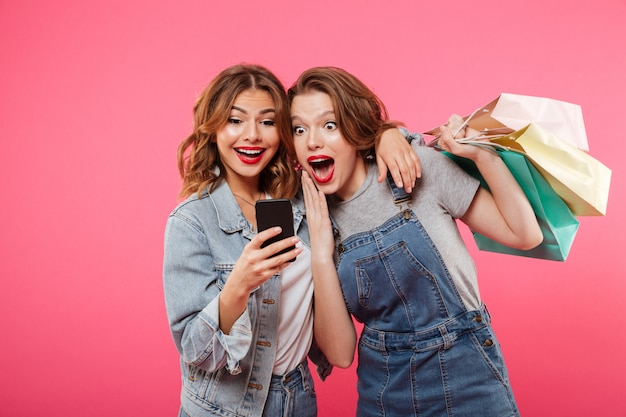 Shocked two women friends holding shopping bags using mobile phone.