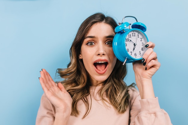 Free photo shocked woman posing with mouth open in morning studio shot of lovely curly girl with clock