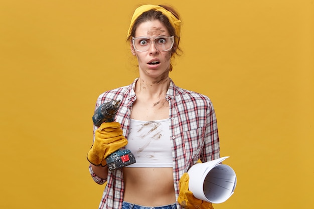 Free photo shocked woman in protective gloves, checkered shirt and yellow headband holding drill and rolled paper having dirty face doing repairing indoors being surprised to see how much she should do