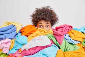 Free photo shocked woman with curly afro hair stares bugged eyes drowned in huge pile of colorful clothing cleans out closet selects clothes for donation or recycling white