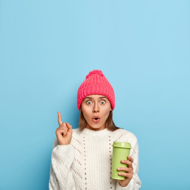 Free photo shocked wondered woman points up with index finger, carries takeaway coffee, scared by something, keeps eyes bugged