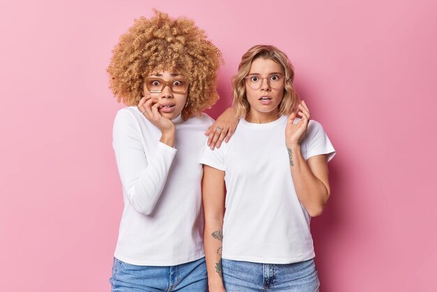 Shocked worried women stareat camera afraid of something see something breathtaking and horrible dressed in casual clothes stand next to each other against pink background Human reactions concept