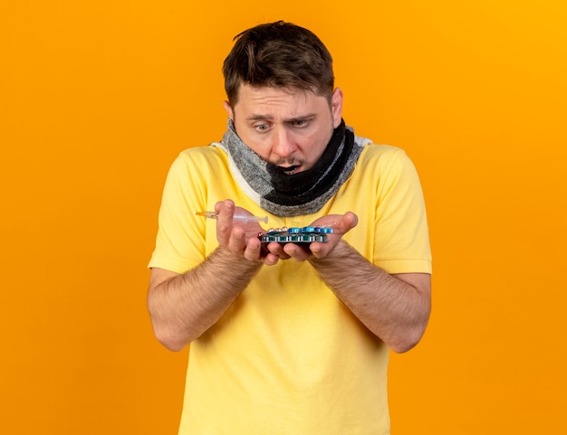 Free photo shocked young blonde ill slavic man covering mouth with scarf holds and looks at packs of medical pills and syringe isolated on orange wall with copy space