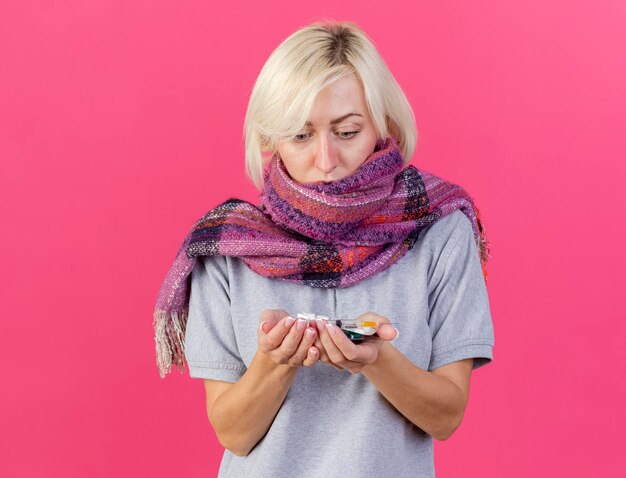 Shocked young blonde ill slavic woman wearing scarf holds and looks at packs of medical pills