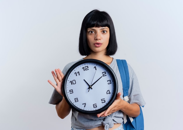 Shocked young brunette caucasian woman wearing backpack holds clock