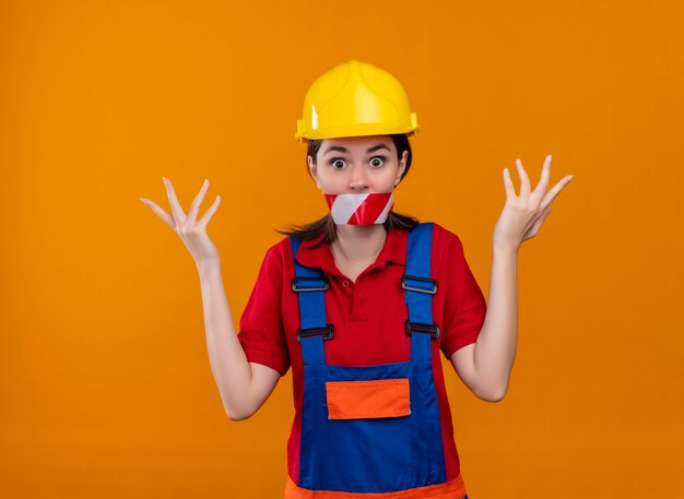 Shocked young builder girl mouth sealed with warning tape raises hands up on isolated orange background