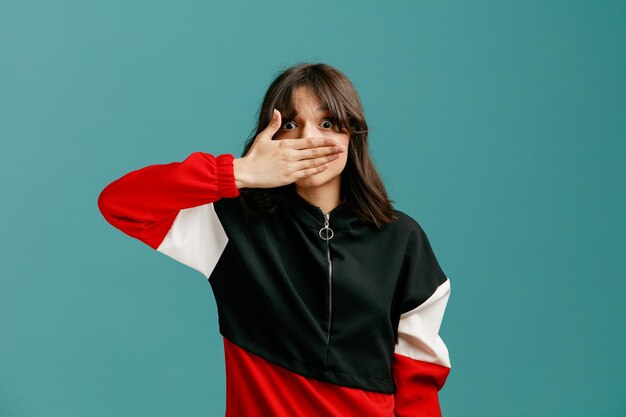 Shocked young caucasian woman looking at camera closing mouth with hand isolated on blue background