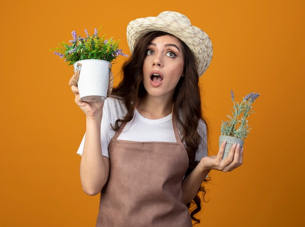 Free photo shocked young female gardener in uniform wearing gardening hat holds flowerpots looking up isolated on orange wall with copy space