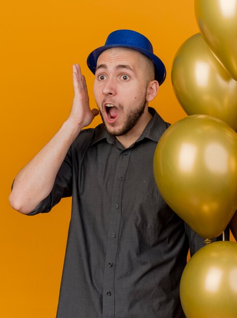 Shocked young handsome slavic party guy wearing party hat holding balloons keeping hand near head looking straight isolated on orange background