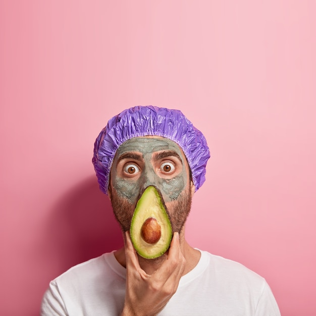 Free photo shocked young man covers nose and mouth with slice of fresh avocado, has eyes popped out, wears clay mask for good effect, waterproof headgear, has beauty procedures at spa center