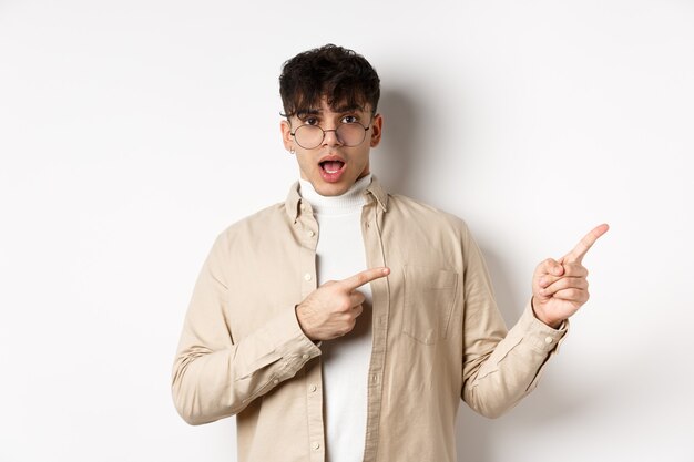 Shocked young man in glasses pointing fingers right at empty space, asking about advertisement, standing on white background