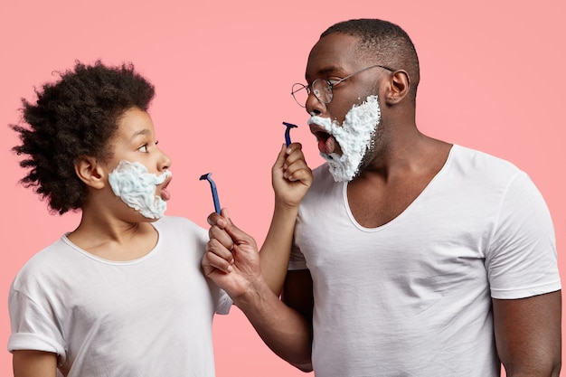 Free Photo shocked young man and his son with shaving foam on faces, keep jaws dropped, hold razors,