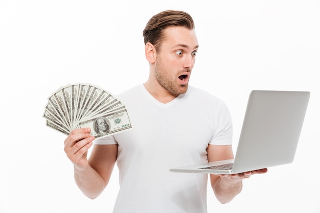 Free photo shocked young man holding money using laptop computer.