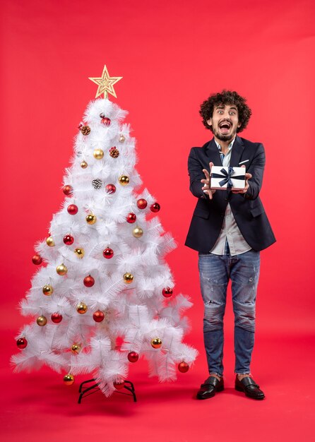 Shocked young man showing his gift standing near decorated white Xmas tree on the right side of red