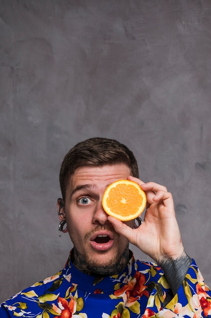 Free photo shocked young man with pierced ears and nose holding and orange slice in front of eyes against grey wall