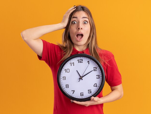 Shocked young pretty woman puts hand on head and holds clock isolated on orange wall