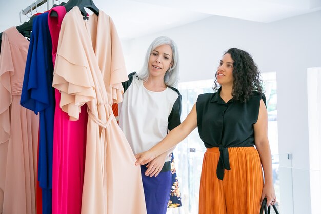 Shop assistant helping customer to choose cloth. Shoppers touching new dress hung on rack. Medium shot. Fashion store or retail concept