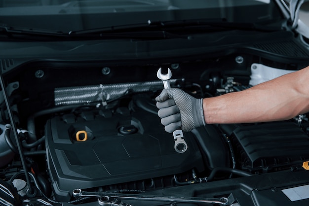 In the shop. Man's hand in glove holds wrench in front of broken automobile