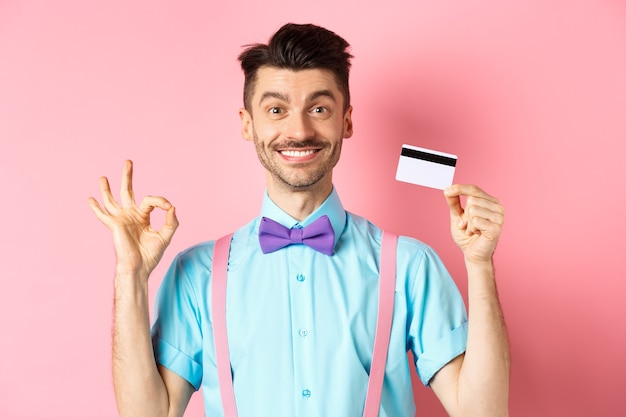 Free Photo shopping concept. smiling handsome male shopper showing ok sign and plastic credit card, buying something, standing satisfied on pink background.