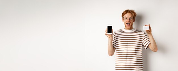 Shopping and finance concept young man showing blank mobile screen and plastic credit card smiling a