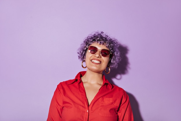 Free photo short-haired curly woman with stylish bright glasses, in red shirt with long sleeve and smiles on isolated lilac.