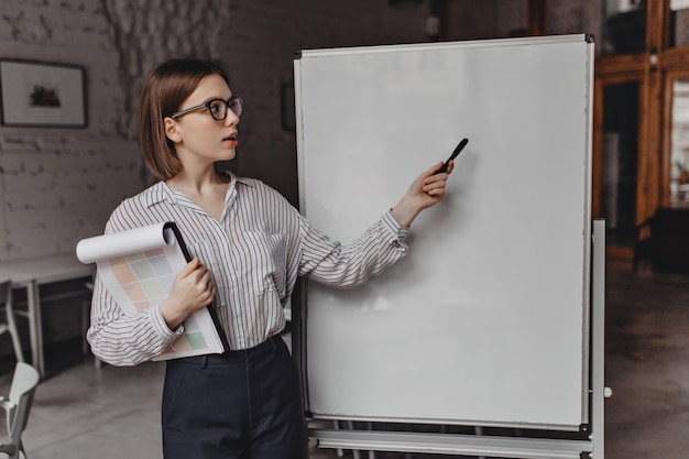 Free Photo short-haired employee in white blouse and black pants shows on office board. portrait of woman with documents telling about plans.