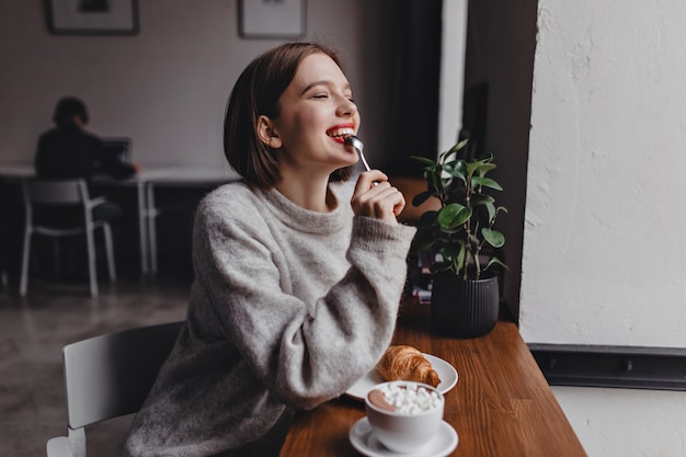 Free Photo short-haired lady in gray sweater bites teaspoon. portrait of girl with red lips sitting in cafe and enjoying croissant and cappuccino.