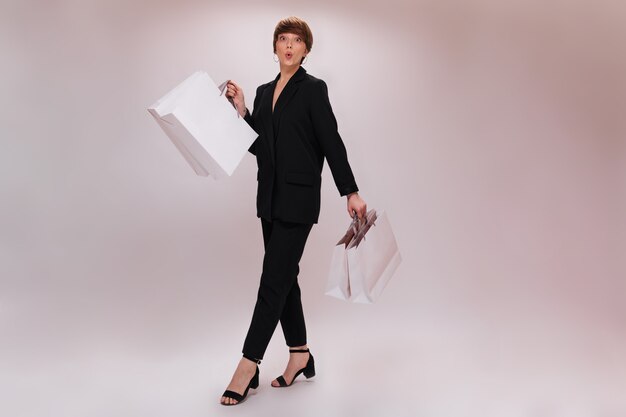 Short haired lady whistles and holds shopping bags. Pretty woman in black suit moves with white packages on isolated backdrop