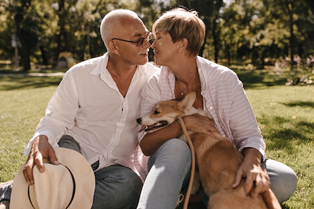 Short haired in striped pink blouse posing with small corgi and kissing man with mustache in white shirt and jeans in park.