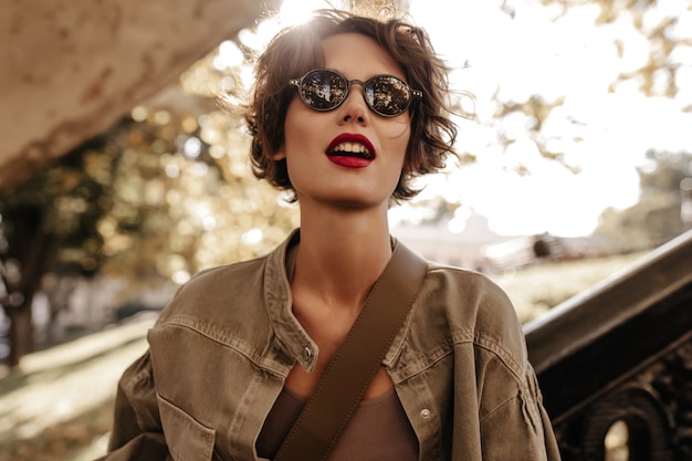 Free photo short-haired woman with black sunglasses in olive jacket looks away outside. wonderful woman with bright lipstick posing outdoors.