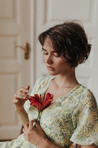 Free photo shorthaired young brunette woman in stylish green and yellow dress looks sad and holds big red flower lady poses near wooden white door