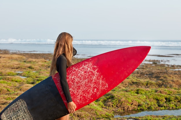 Shot of active sporty woman carries red surfboard with waxed surface