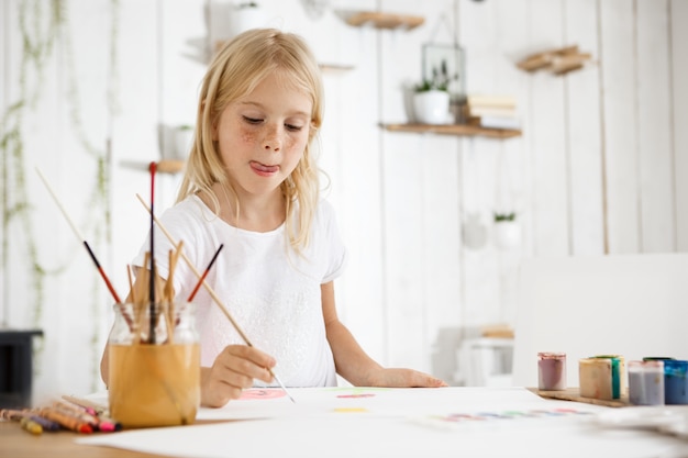 Free photo shot of adorable blonde girl with freckles biting her tongue because of inspiration while painting. girl with blond hair sitting at the room filled with morning light and wearing white clothes.