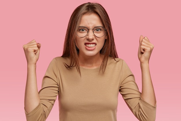 Shot of aggressive woman raises hands clenched in fists, argues with neighbours, looks intense and irritated, expresses irritation, wears beige sweater, isolated over pink wall. Crazy girl