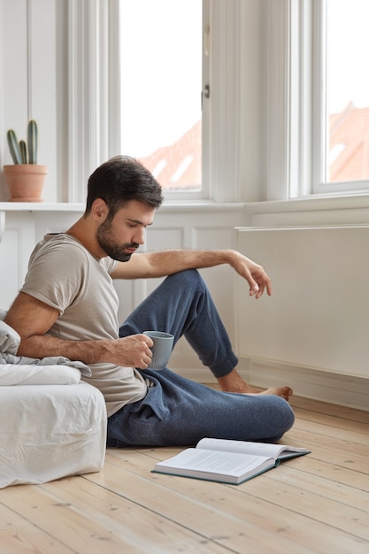 Shot of attractive clever man enjoys reading book at home sits at floor near bed, drinks fresh hot beverage, likes novel, feels inspired and realxed, enjoys calm atmosphere. Literature develops us