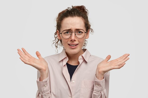 Free photo shot of attractive young female with hesitant expression, shruggs shoulders and keeps palms raised, has clueless look, isolated over white wall. puzzlement and doubt concept.