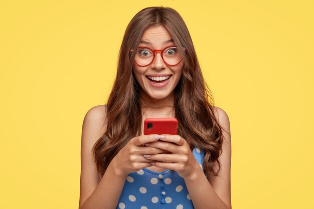 Shot of cheerful female model wears glasses, holds cell phone, takes pictures, talks via video message, has nice friendly talk, dressed in fashionable outfit, isolated over yellow wall