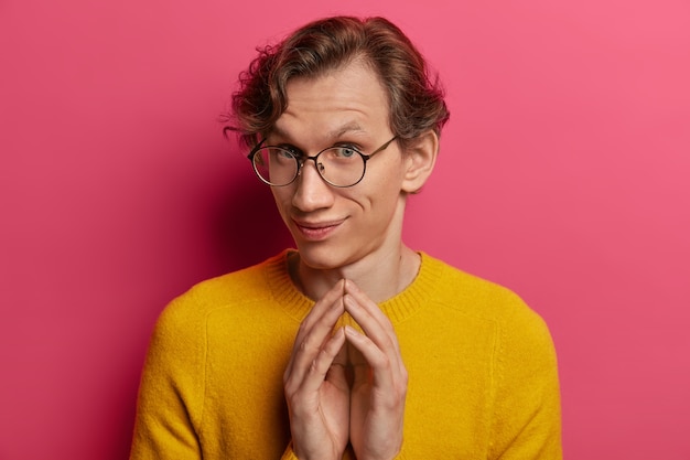 Free photo shot of curious young caucasian man steepls fingers, looks with intention to do something, wears round spectacles and yellow jumper, has some plans in mind, isolated over pink wall