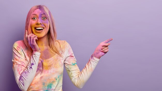 Shot of glad blonde woman splattered with multicolored powder, shows place where holi color festival is, expresses positive emotions, impressed by festive events in India, likes coloured throw