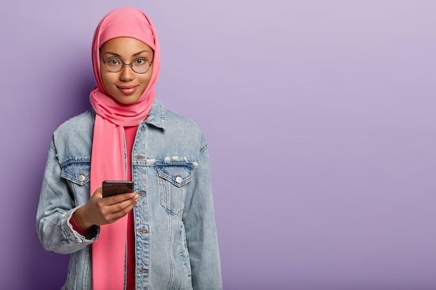 Free photo shot of good looking dark skinned woman with appealing look wears pink hijab and denim coat, holds modern cell phone, waits for important call, stands over purple wall with blank space