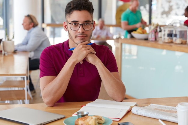 Shot of handsome serious mixed race young man writer holds pen, makes list to do in notepad, drinks coffee, spends lunch time in coffee shop alone, busy with work.