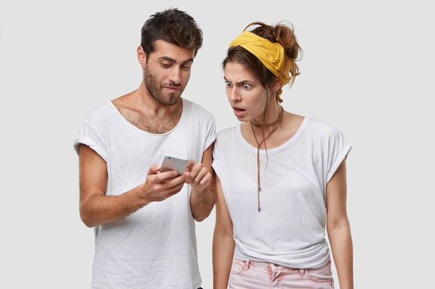 shot of handsome unshaven man shows some web site news to his European woman, holds cell phone, stands over white wall, enjoy high speed internet on electronic device