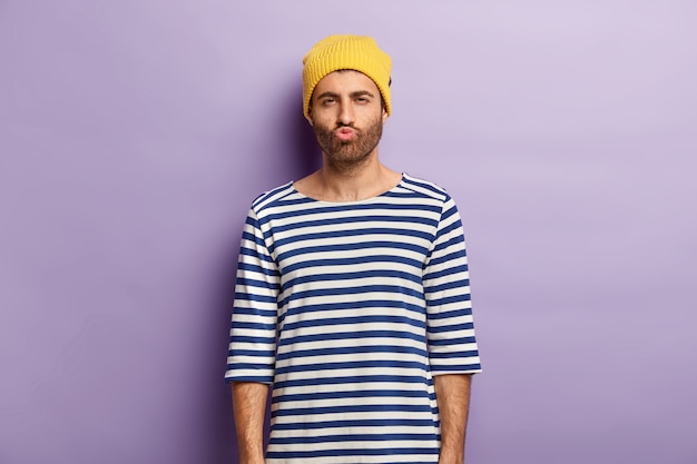 Free Photo shot of handsome youngster with bristle pouts lips and looks mysteriously, being dissatisfied with something, wears yellow hat and striped sailor jumper